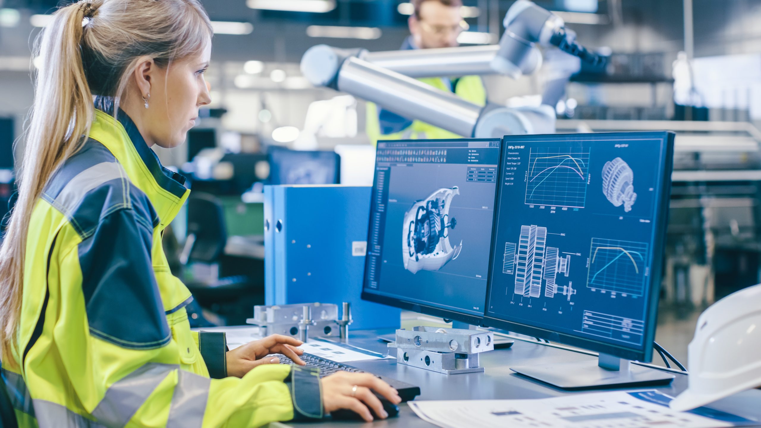 At the Factory: Female Mechanical Engineer Designs 3D Engine on Her Personal Computer, Male Automation Engineer Uses Laptop for Programming Robotic Arm. At the Factory: Female Mechanical Engineer Designs 3D Engine on Her Personal Computer, Male Automation Engineer Uses Laptop for Programming Robotic Arm.