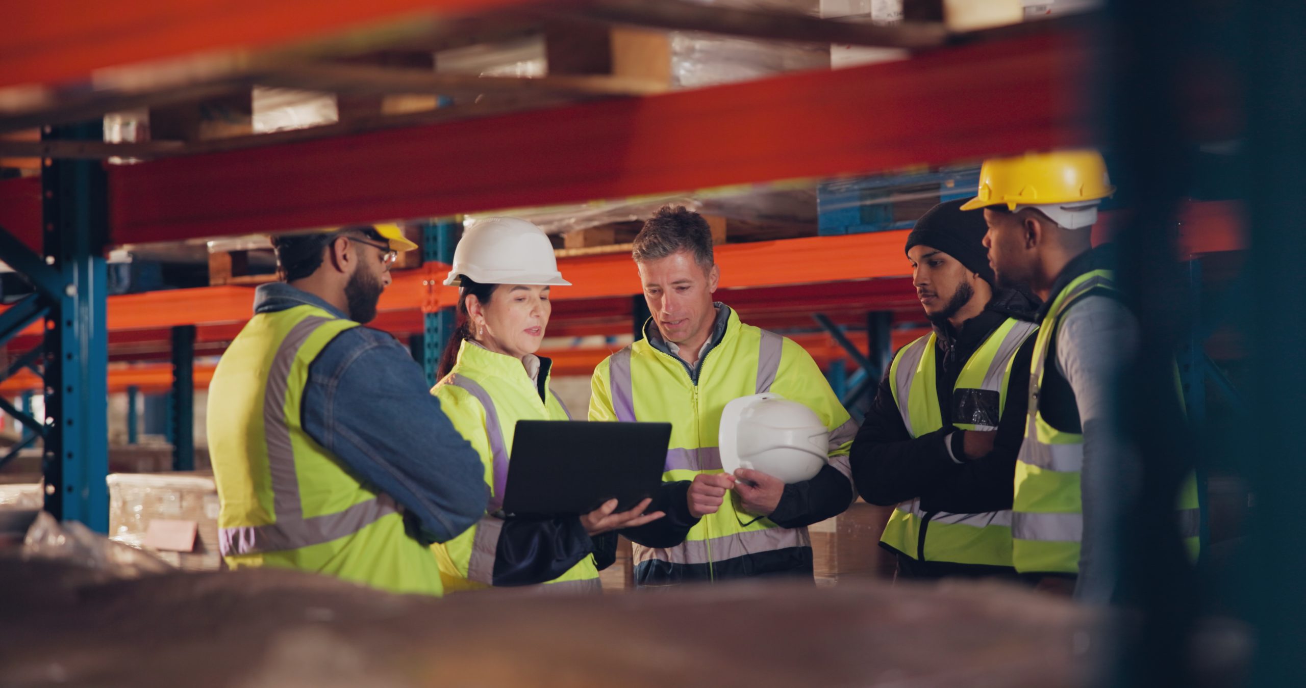 Industrial Team Discussing Safety Performance Five industrial team members in high-visibility clothing reviewing information on a laptop inside a warehouse.
